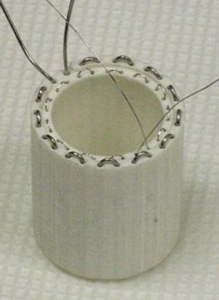 A white, cylindrical 3D-printed part with a metal wire coil threaded through small holes along the top rim, shown against a textured white background in "csl_nasa_rover_heater_lo-res_prodways_printers.jpeg